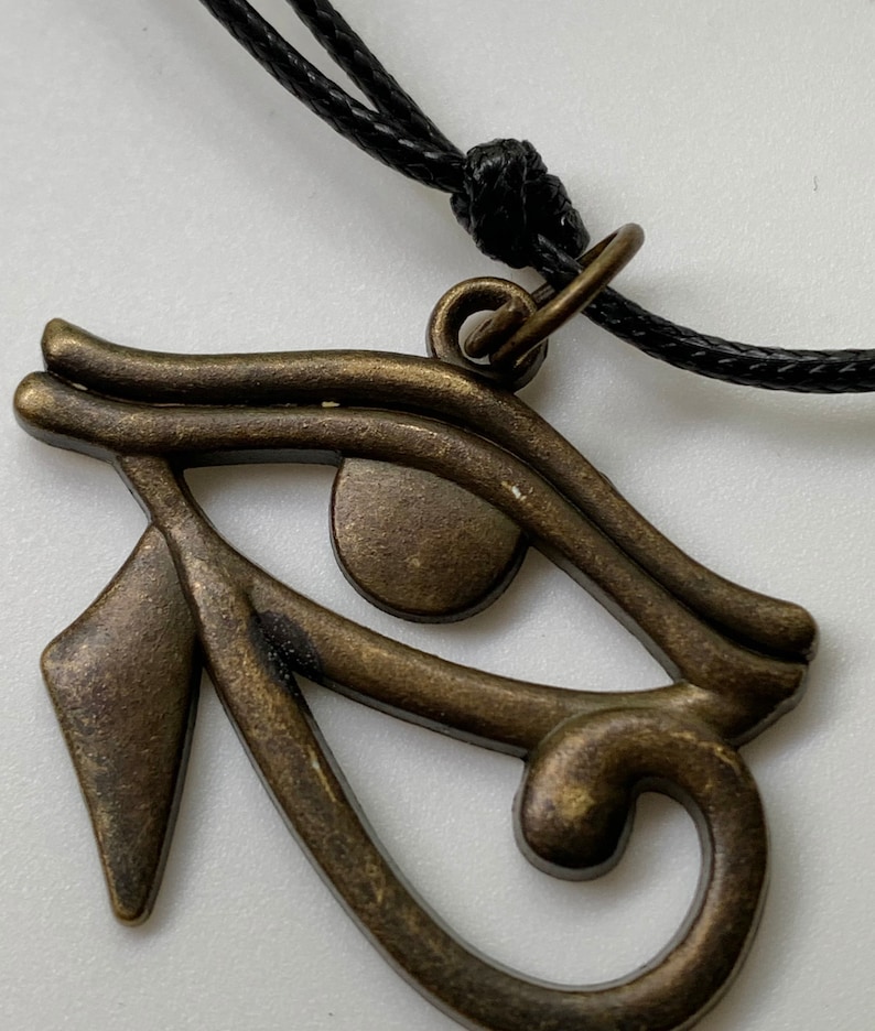 May include: A bronze-colored Eye of Horus pendant on a black cord necklace. The pendant is detailed with intricate lines and a circular eye.