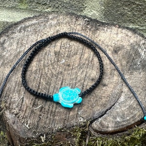 Puede incluir: Un brazalete de cordón trenzado negro con un dije de tortuga turquesa. El brazalete tiene lazos ajustables en cada extremo.