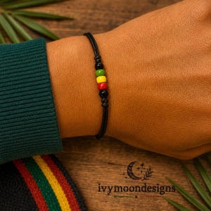 Puede incluir: Una pulsera de cordón negro con cinco cuentas pequeñas en disposición vertical. Las cuentas son de color negro, verde, amarillo, rojo y negro, aseguradas con nudos en un cordón negro fino. La pulsera se lleva en la muñeca, mostrando la secuencia.