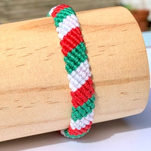 Könnte beinhalten: Ein grünes, weißes und rotes Freundschaftsarmband mit einem diagonalen Muster. Das Armband ist aus geflochtener Schnur gefertigt und wird auf einer Holzoberfläche präsentiert.