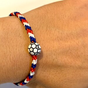 May include: A red, white, and blue braided bracelet with a soccer ball charm. The bracelet is on a person's wrist.
