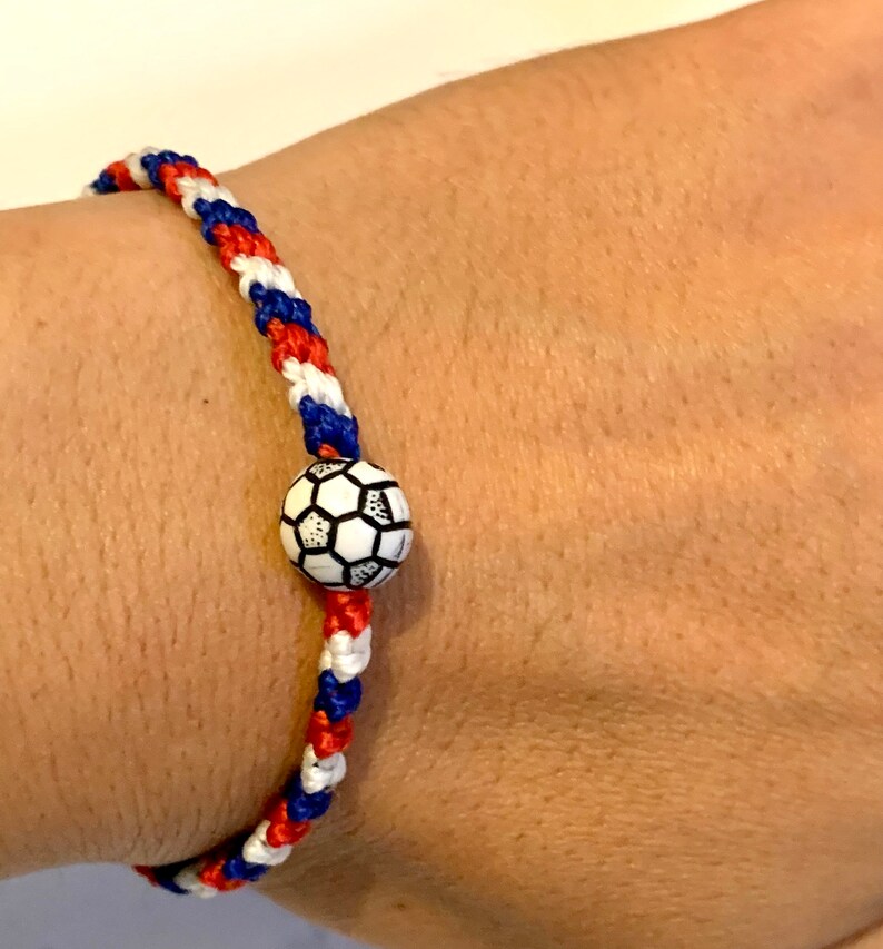 May include: A red, white, and blue braided bracelet with a soccer ball charm. The bracelet is on a person's wrist.