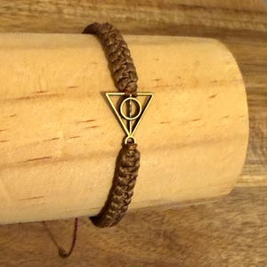 Op de afbeelding: Een bruine macramé armband met een goudkleurige driehoek bedel. De bedel heeft een cirkel in de driehoek. De armband wordt getoond op een houten oppervlak.