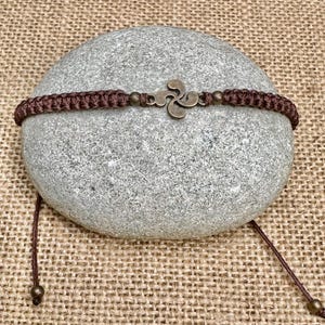 May include: A brown macrame bracelet with a bronze-colored Basque cross charm. The bracelet is displayed on a gray stone, set against a textured, tan background. The bracelet has adjustable cords with bronze-colored beads.
