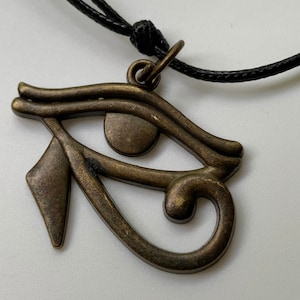 May include: A gold-toned Eye of Horus pendant necklace with a black cord. The pendant is a detailed representation of the ancient Egyptian symbol.