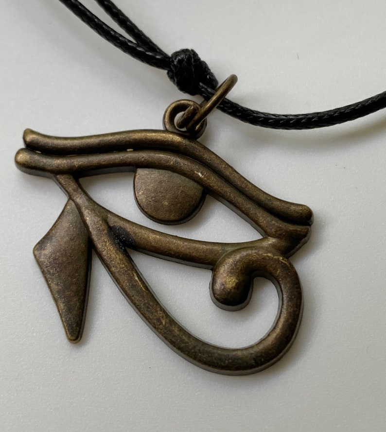 May include: A gold-toned Eye of Horus pendant necklace with a black cord. The pendant is a detailed representation of the ancient Egyptian symbol.