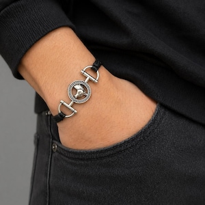 Könnte beinhalten: Ein schwarzes Kordelarmband mit silberfarbenen Metallakzenten. Das Armband hat einen runden Anhänger mit einem Pferdekopf-Design. Das Armband wird mit zwei D-förmigen Verschlüssen befestigt.