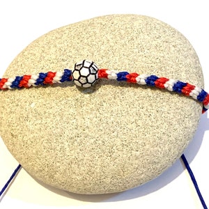 May include: A red, white, and blue braided friendship bracelet with a white soccer ball charm. The bracelet is tied around a grey rock.