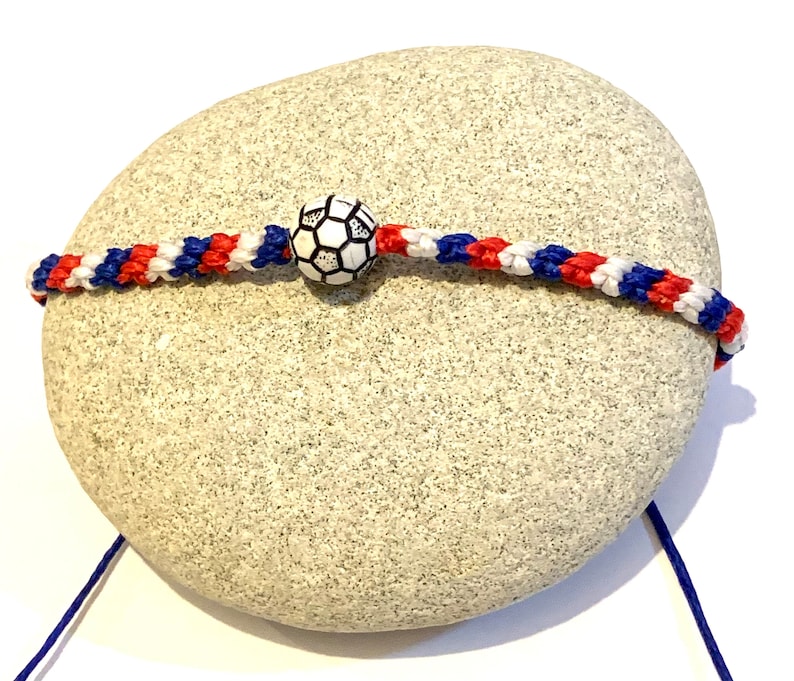 May include: A red, white, and blue braided friendship bracelet with a white soccer ball charm. The bracelet is tied around a grey rock.