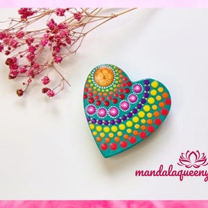Può includere: Un ornamento di legno a forma di cuore color turchese con un design di mandala colorato dipinto su di esso. Il mandala presenta una varietà di colori, tra cui rosso, arancione, giallo, verde, blu e viola. L'ornamento è circondato da uno sfondo bianco.