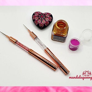 Può includere: Due strumenti di punteggio in oro rosa con accenti di cristallo trasparente e rosa. Gli strumenti sono su una superficie bianca con una pittura di roccia a forma di cuore rosa, un flacone di vernice dorata e un flacone di vernice rosa.