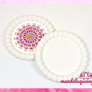 Pode incluir: Dois  copos de cerâmica branca redondos, um com um design de mandala rosa e dourado. Os copos têm uma borda de contas.