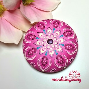 Könnte beinhalten: Ein handbemalter, runder, rosa Mandala-Stein mit einem detaillierten Blumendesign in Rosa-, Weiß- und Türkistönen. Der Stein hat einen zentralen lila Edelstein und den Text "mandalaqueeny" in Rosa.