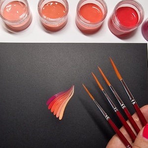 May include: A collection of art supplies including small jars of various shades of paint, a black surface with a painted design, and several fine-tipped paint brushes with red handles.