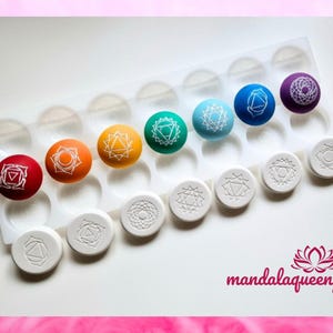 Könnte beinhalten: Eine Silikonform mit 12 runden Vertiefungen, jede mit einem andersfarbigen Chakra-Symbol. Die Symbole sind weiß und die Form ist weiß. Die Form befindet sich auf einer weißen Oberfläche. Der Text "mandalaqueen" ist in der rechten unteren Ecke des Bildes sichtbar.