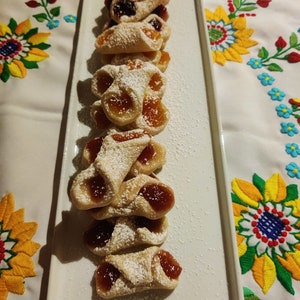 Pu&ograve; includere: Un piatto bianco con una fila di biscotti fatti in casa a forma di mezzaluna, ripieni di marmellata e spolverati di zucchero a velo.
