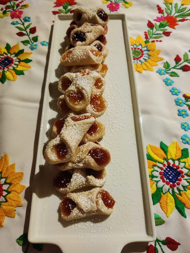 Pu&ograve; includere: Un piatto bianco con una fila di biscotti fatti in casa a forma di mezzaluna, ripieni di marmellata e spolverati di zucchero a velo.