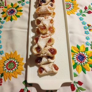 Pu&ograve; includere: Un piatto rettangolare bianco con una fila di pasticcini fatti in casa. I pasticcini sono ripieni di marmellata e ricoperti di zucchero a velo. Il piatto &egrave; su una tovaglia bianca con fiori ricamati colorati.