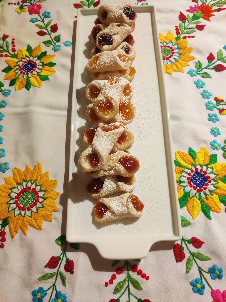 Pu&ograve; includere: Un piatto rettangolare bianco con una fila di pasticcini fatti in casa. I pasticcini sono ripieni di marmellata e ricoperti di zucchero a velo. Il piatto &egrave; su una tovaglia bianca con fiori ricamati colorati.