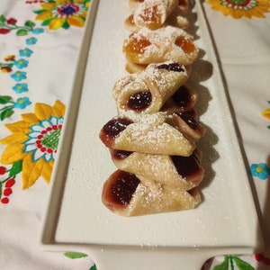 Pu&ograve; includere: Un piatto bianco con una pila di pasticcini fatti in casa. I pasticcini sono ripieni di marmellata e ricoperti di zucchero a velo.