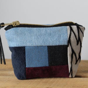 Puede incluir: Un pequeño monedero de patchwork con cremallera negra y tirador de cuero negro. El bolso está hecho de varias telas en tonos azules, negros y burdeos. La cremallera es dorada. El bolso está sobre una superficie de madera.