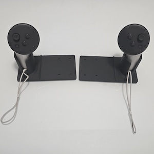May include: Two black VR controller mounts. Each mount has a circular top with buttons, a curved support, and a rectangular base with mounting holes. White lanyards hang from the base.