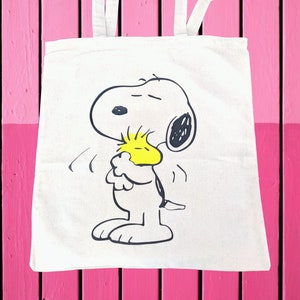 May include: Off-white canvas tote bag featuring a black and white illustration of Snoopy hugging Woodstock. The bag has two white handles and is set against a pink wooden background. The cartoon characters are in a joyful embrace.