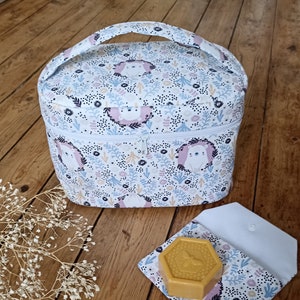 Op de afbeelding: Een witte toilettas van stof met een ritssluiting en een handvat. De tas heeft een bloemenpatroon met kleine witte beren. De tas ligt op een houten oppervlak met een witte stoffen tas en een gele zeep.