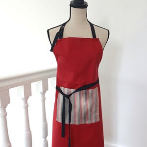 Peut inclure: Un tablier rouge avec une sangle de cou et une ceinture noires. Le tablier a une poche avec un motif à rayures de lignes verticales rouges, noires et crème. Le tablier est présenté sur un mannequin.