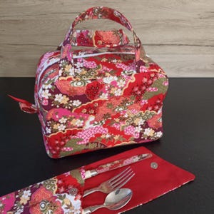 Peut inclure: Un sac à déjeuner en tissu rouge avec un motif floral et géométrique, doté d'une poignée supérieure et d'une fermeture éclair. À côté se trouve une pochette à couverts assortie contenant une fourchette, une cuillère et un couteau en argent.