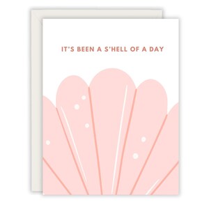 May include: A pink and white greeting card with the text "It's been a s'hell of a day". The card features a pink seashell illustration.