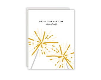 Könnte beinhalten: Eine weiße Grußkarte mit zwei Illustrationen von goldenen und hellgelben Wunderkerzen im unteren Bereich. Der Text oben lautet I HOPE YOUR NEW YEAR is a blast in schwarzer, serifenloser und kursiver Schrift. Das Design ist schlicht.