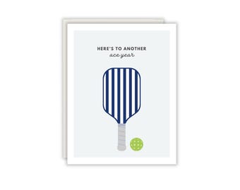 Könnte beinhalten: Eine weiße Grußkarte mit einer dunkelblau-weiß gestreiften Pickleball-Schläger-Grafik, einem grauen Griff und einem hellgrünen Pickleball. Der Text oben lautet "HERE'S TO ANOTHER ace year" in dunkelgrauer, serifenloser und kursiver Schrift.