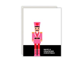 Könnte beinhalten: Eine Grußkarte mit einer Illustration eines rosa Nussknackers mit einem roten Herz auf Hut und Stiefeln. Der Nussknacker trägt eine rosa Uniform mit roten Streifen und einem schwarzen Gürtel. Der Text "HAVE A CRACKING CHRISTMAS" steht in Weiß auf schwarzem Grund.