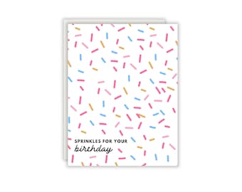 Könnte beinhalten: Eine rechteckige Grußkarte mit weißem Hintergrund und einem Muster aus kleinen, verstreuten, strichförmigen Streuseln in Rosa, Hellblau und Gold. Der Text "SPRINKLES FOR YOUR birthday" ist unten links in Schwarz aufgedruckt.