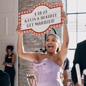 May include: A woman in a lavender dress holds a sign that reads "4.18.25 KEVIN & DESIREE GET MARRIED!" The sign has a red and white border with light bulbs. The woman is smiling and appears to be celebrating.