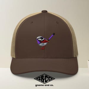May include: A brown trucker cap with a beige mesh back and a brown peak. The cap has an embroidered bird with a white head, purple details, and red feet. The logo "G&CD gnome and co." is at the bottom.