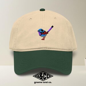 Op de afbeelding: Beige en donkergroene baseballcap met een geborduurd vogelontwerp. De vogel is blauw, paars en rood. De cap heeft een donkergroen vizier en een kleine groene knop. Het logo "G&CD gnome and co." staat onderaan.
