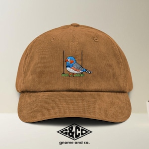 Könnte beinhalten: Braune Cord-Baseballkappe mit einem gestickten Vogeldesign. Der Vogel ist blau, orange und grau und sitzt auf einem grünen Ast. Die Kappe hat einen gebogenen Schirm und ein klassisches Sechs-Panel-Design. Das Markenlogo befindet sich unten.