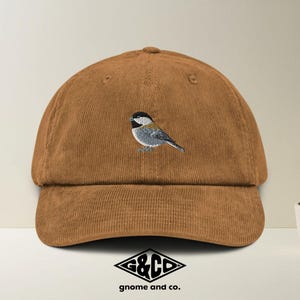 Peut inclure: Casquette de baseball en velours côtelé marron avec un motif d'oiseau brodé. L'oiseau est gris, noir et blanc. La casquette a une visière incurvée et un logo "G&CO gnome and co." en bas.