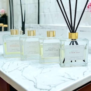 May include: Four glass bottles with gold lids and black reeds, filled with scented oil. The bottles are labeled with different scents: Watermint Clementine, Palo Santo, Bamboo Coconut Lime, and A New Day. The bottles are on a white marble countertop.