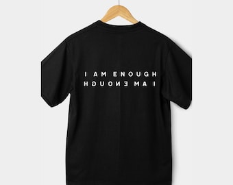 May include: Black t-shirt with the words "I AM ENOUGH" printed in white, with the same words mirrored below. The shirt is hanging on a wooden hanger.