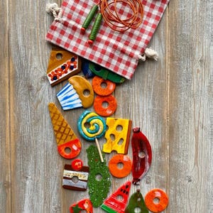 May include: A red and white checkered bag with a jump rope and a variety of colorful wooden food-shaped toys. The toys include a cupcake, a lollipop, a slice of watermelon, a slice of cheese, a strawberry, a slice of cake, a cone, a donut, and a pomegranate.