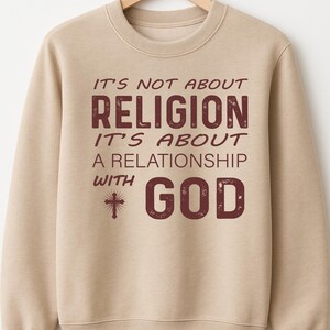 May include: A beige sweatshirt with the text "IT'S NOT ABOUT RELIGION IT'S ABOUT A RELATIONSHIP WITH GOD" in maroon lettering. The sweatshirt is hanging on a wooden hanger.