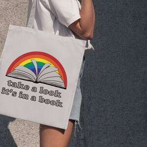May include: A white tote bag with a rainbow and an open book design. The text on the bag reads "take a look it's in a book".