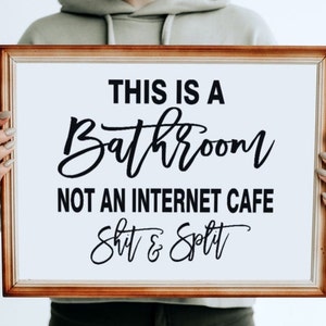 May include: A framed sign with the text "THIS IS A BATHROOM NOT AN INTERNET CAFE Shit & Split" in black script and block letters. The frame is a warm brown color, and the background is white.