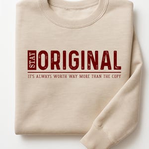 May include: A beige sweatshirt with the words "STAY ORIGINAL" in burgundy block letters. Below the text is the phrase "IT'S ALWAYS WORTH WAY MORE THAN THE COPY". The sweatshirt is folded.