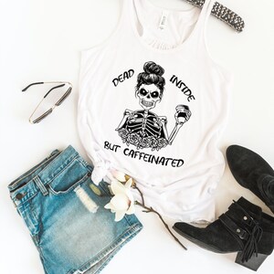 May include: White tank top with a black and white illustration of a skeleton holding a coffee cup. The text "Dead Inside But Caffeinated" is printed on the shirt.