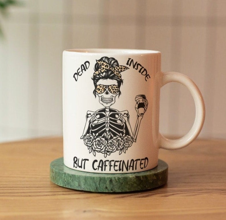May include: White ceramic coffee mug with a black and white illustration of a skeleton wearing sunglasses and a leopard print headband. The text "Dead Inside But Caffeinated" is printed on the mug.