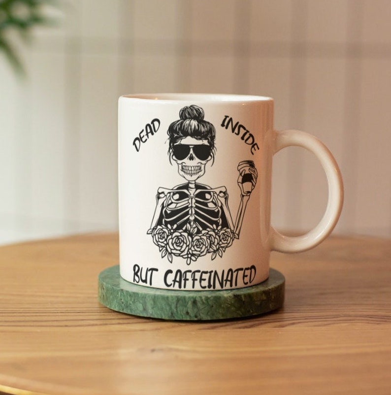 May include: White ceramic coffee mug with a black and white illustration of a skeleton holding a coffee cup. The text "Dead Inside But Caffeinated" is printed on the mug. The mug is sitting on a green marble coaster.